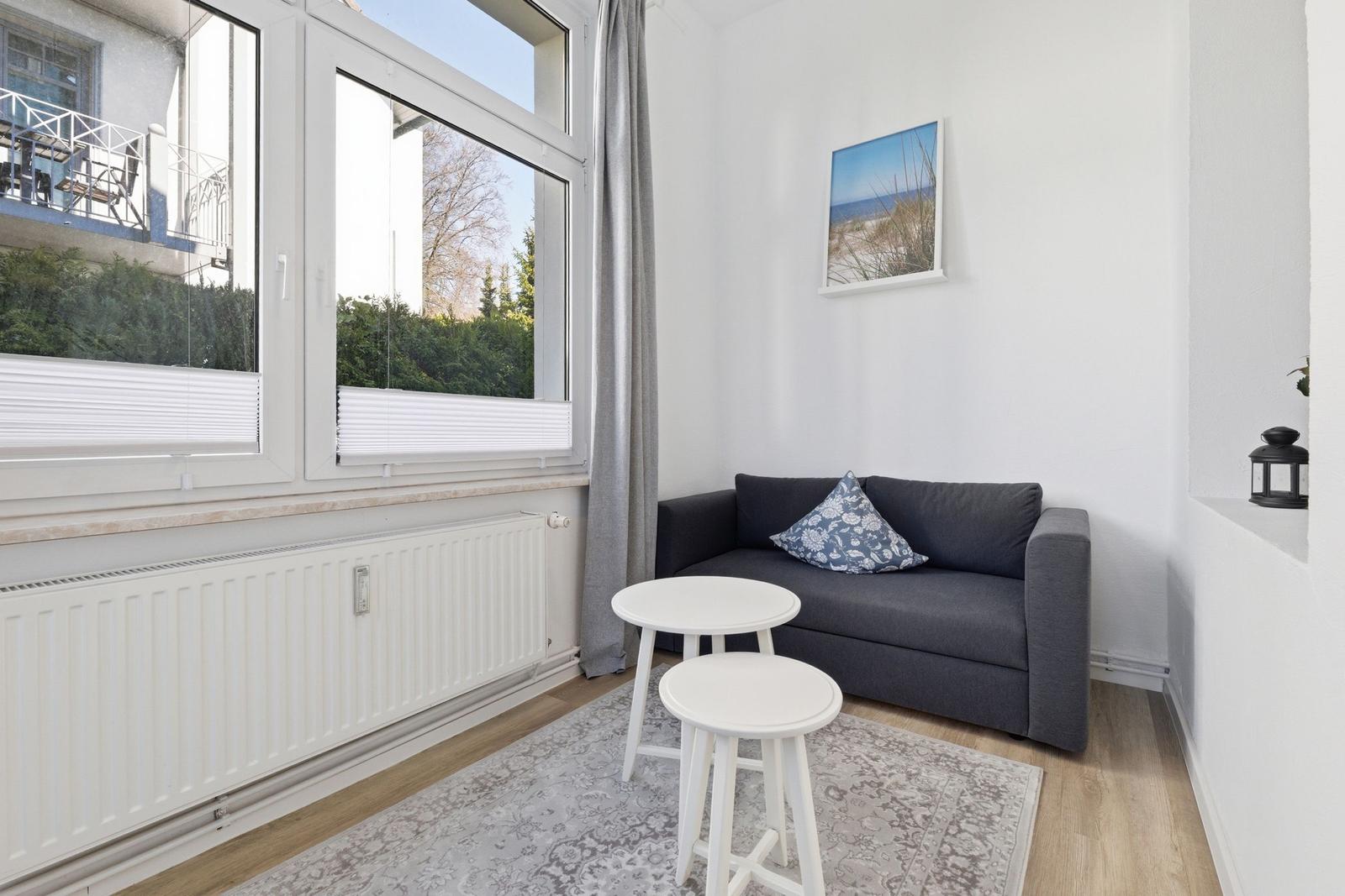 Ein kleiner Wohnbereich mit grauem Sofa, zwei weißen Tischen und großem Fenster.