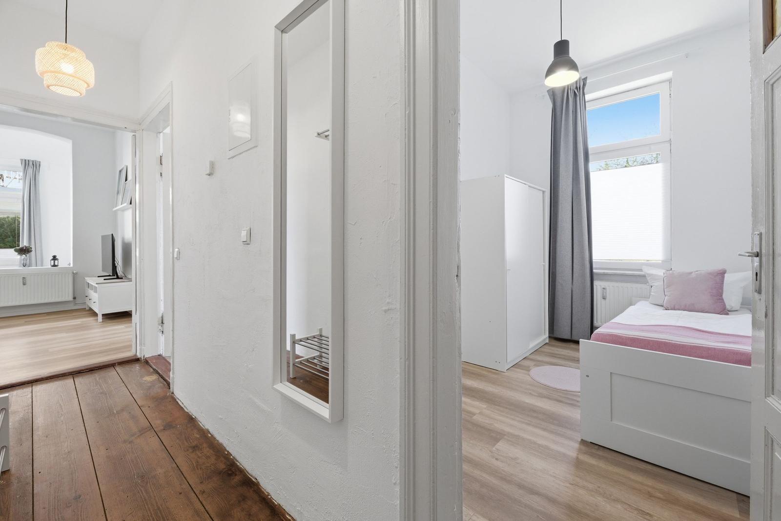 Schlafzimmer mit Bett, Kleiderschrank und Fenster. Flur mit Spiegel und Holzboden. Wohnzimmer im Hintergrund.