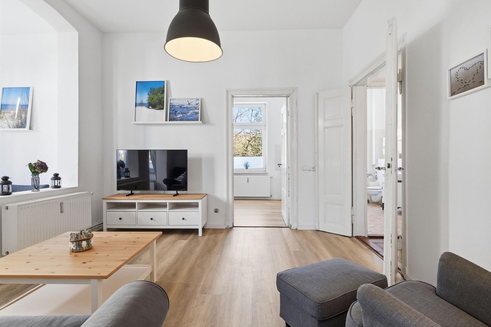 Wohnzimmer mit Fernseher, Holztisch und grauem Sofa. Durchgänge zu Schlaf- und Badezimmer.