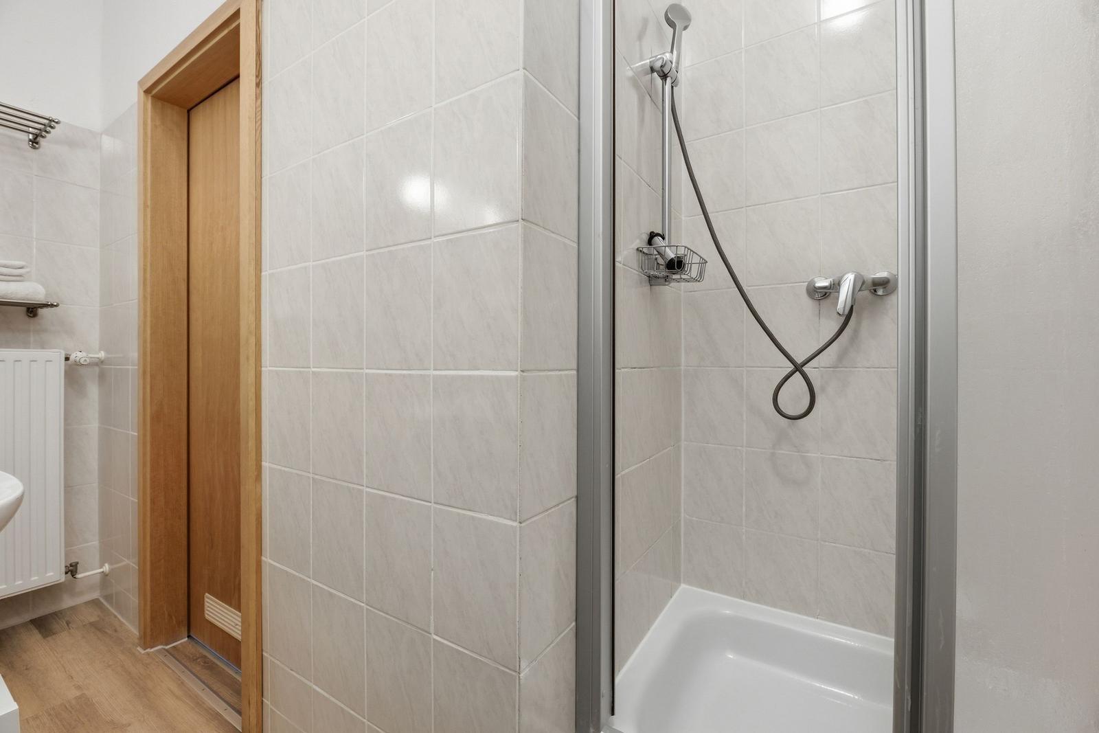 Bath with shower, tiled wall, and wooden door. Hand shower and shower rail visible.