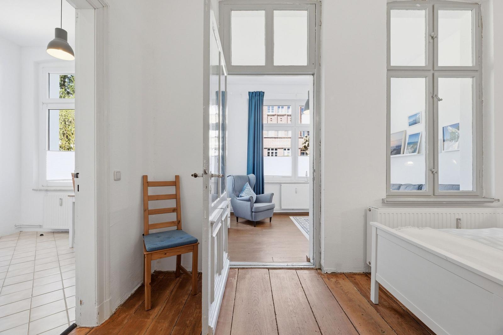 Bedroom with wooden floor and white bed frame. Door leads to living area with armchair.