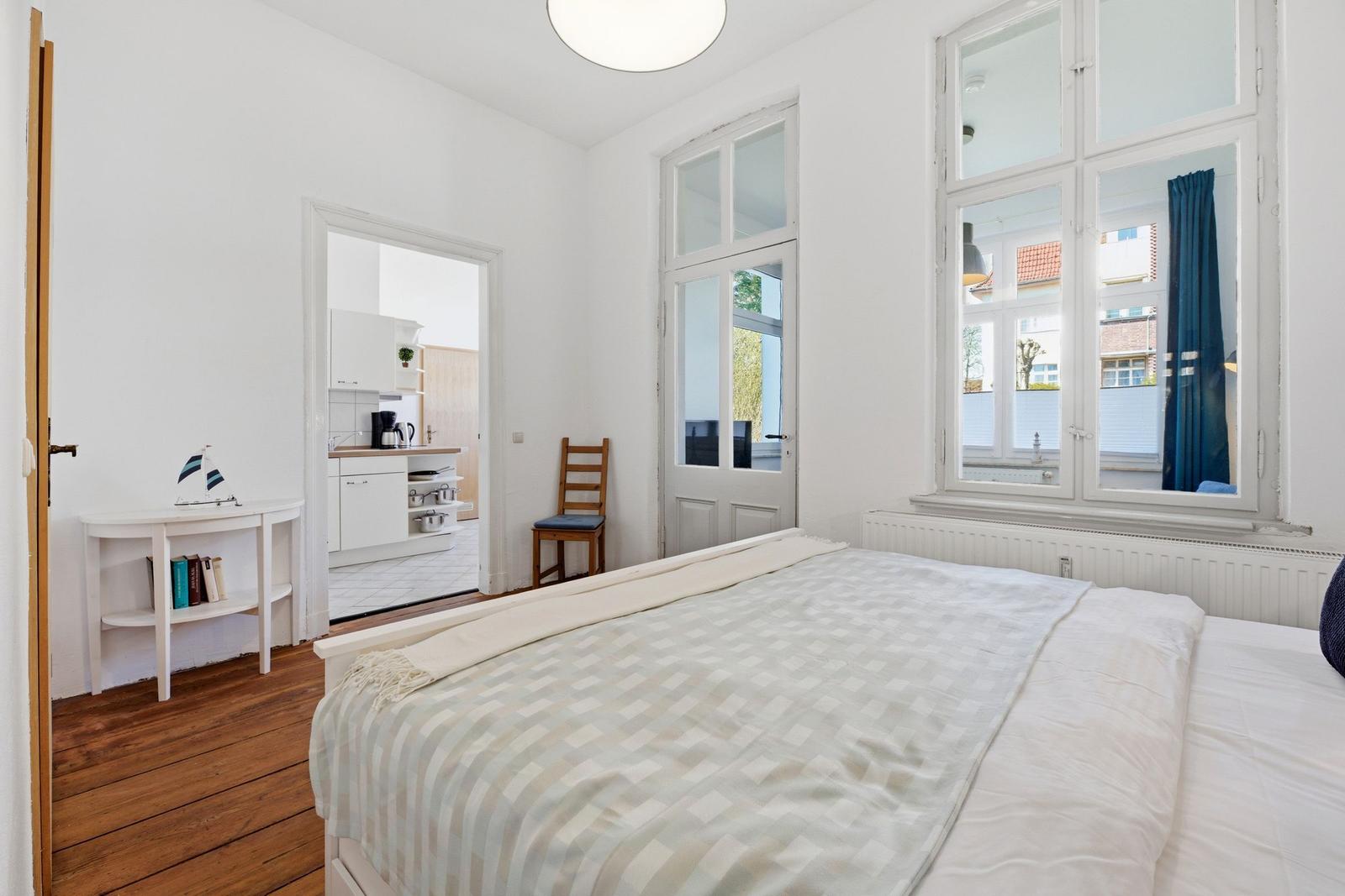 Bedroom with bed, wooden floor, and window. Kitchen visible in background.