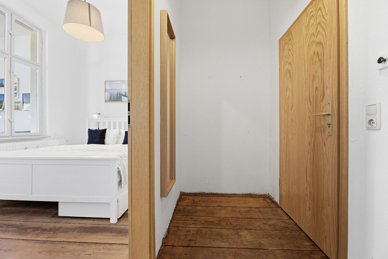 Bedroom with white bed and wooden floor. Door and mirror on the wall.