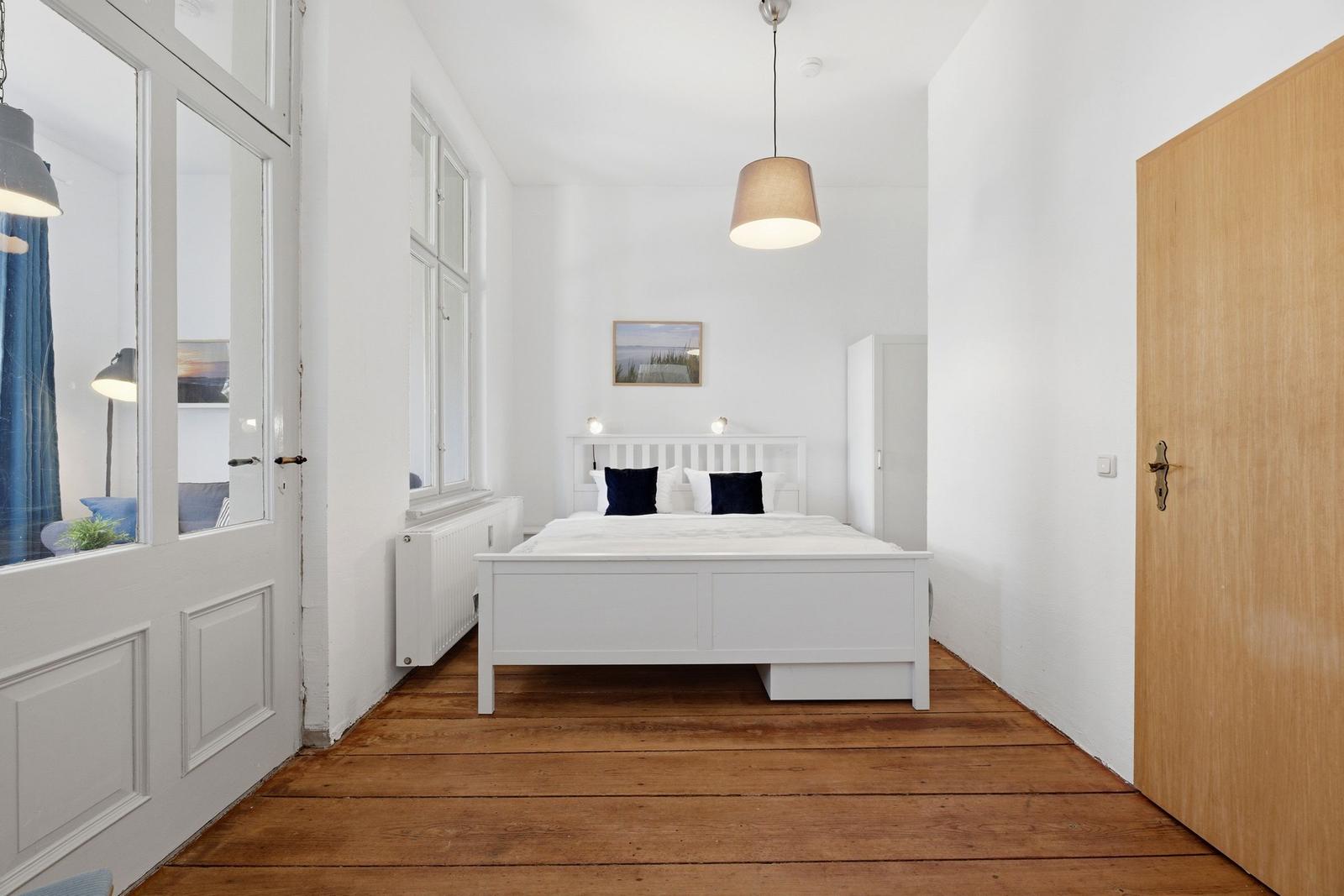 Bedroom with white bed, wooden floor, and window. Pendant lamp and wall picture visible.