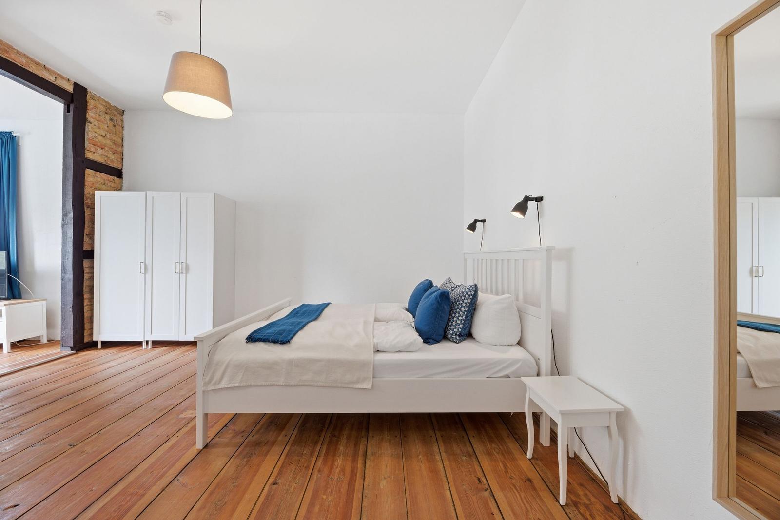 Schlafzimmer mit weißem Bett, Holzboden und Spiegel. An der Wand hängen zwei Lampen.