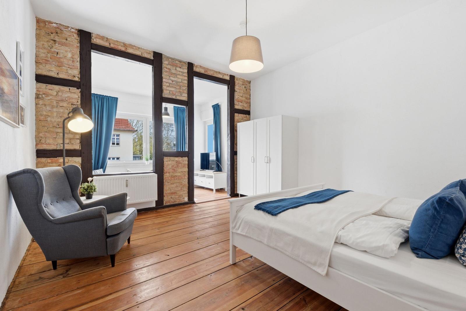 Schlafzimmer mit Bett, Stuhl, Kleiderschrank und Holzboden. Blick auf Wohnbereich mit Fenstern.