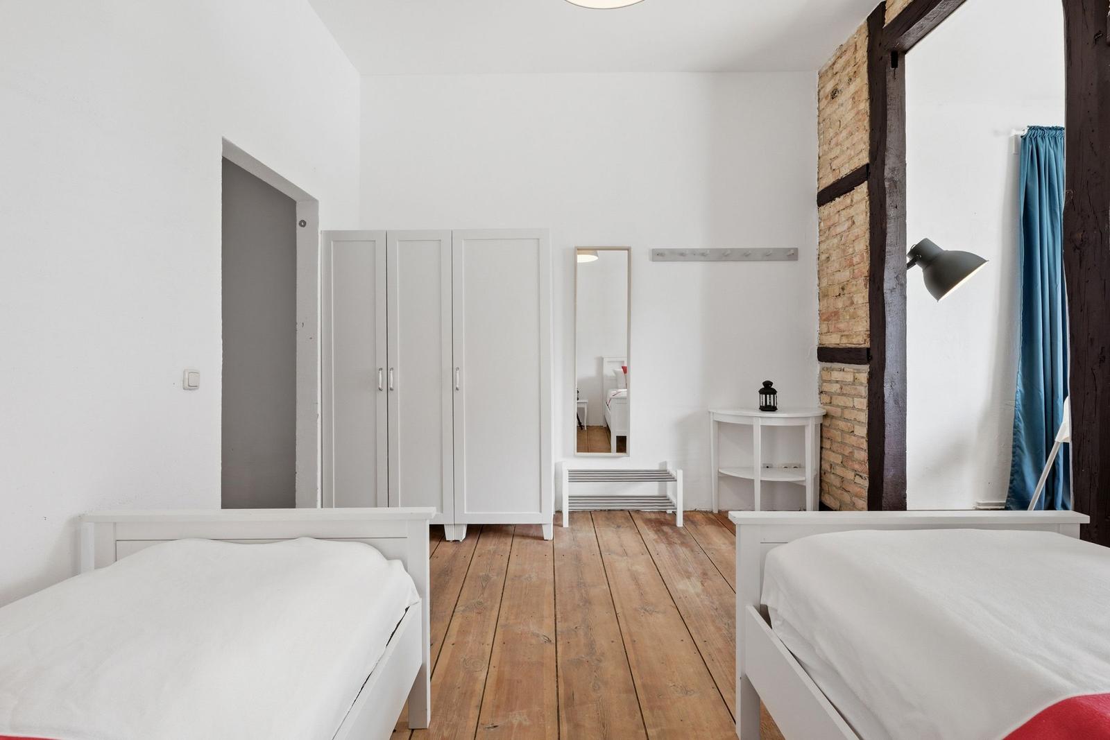 Zwei Betten, weiße Kleidung, Holzboden, Spiegel, Schrank und Lampe.