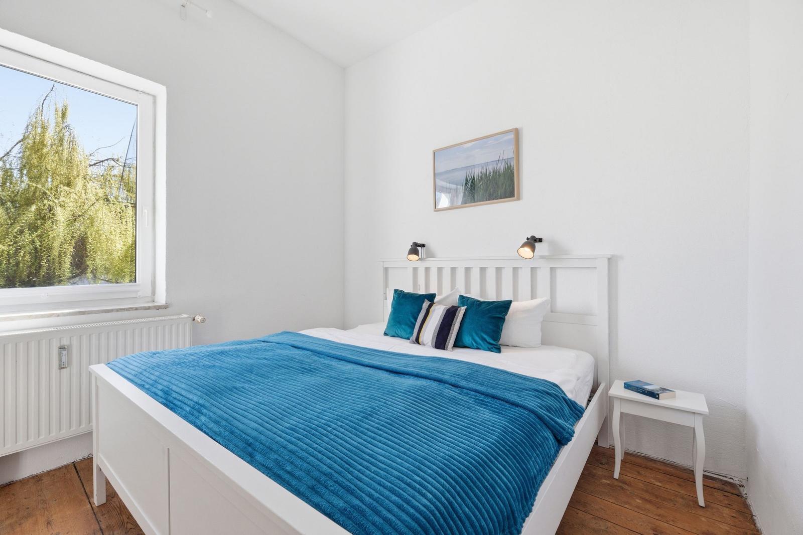 A bedroom with a white bed, blue blanket, and window with view of trees.