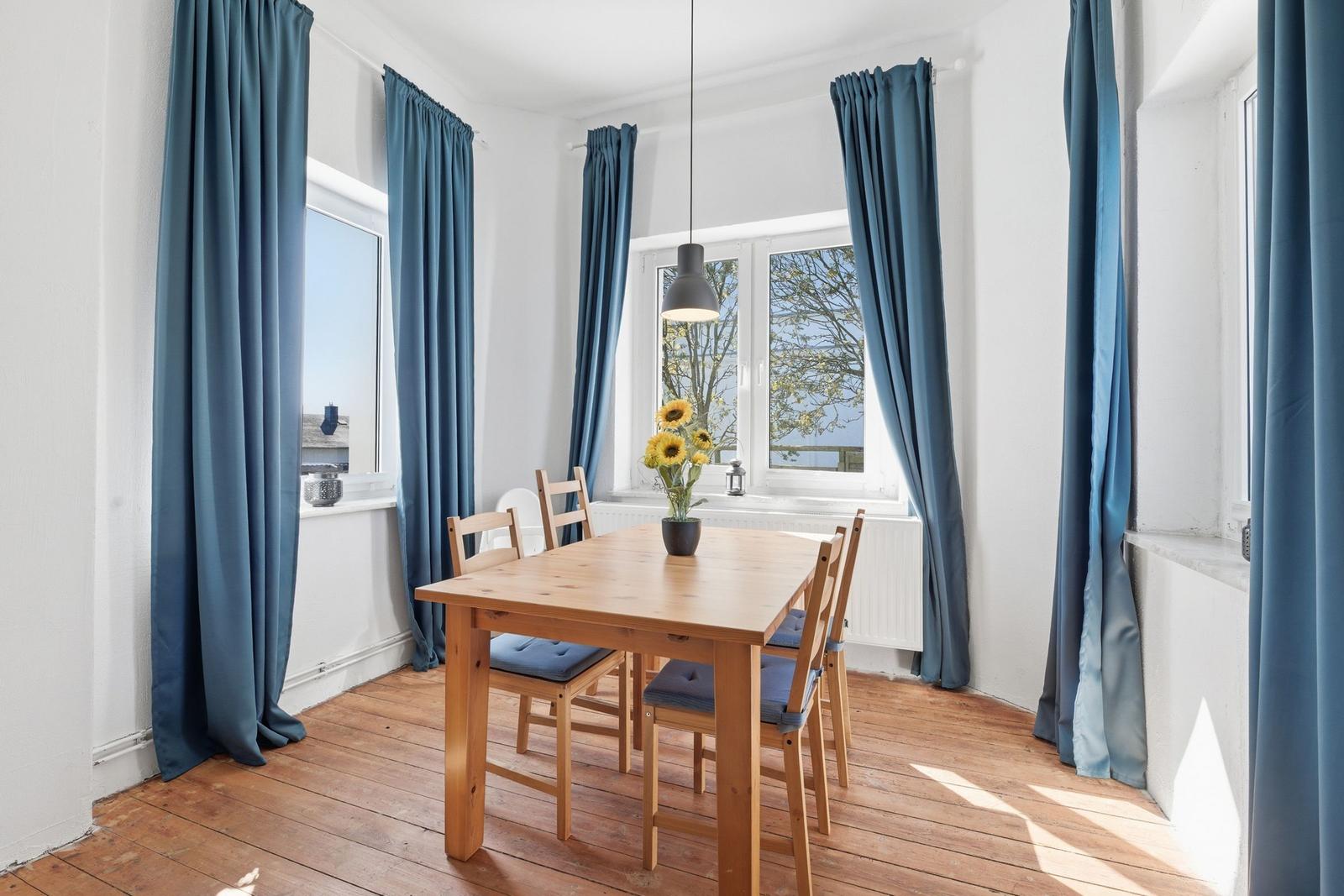Dining room with wooden table, chairs, and blue curtains. Sunflowers on table. Wooden floor.
