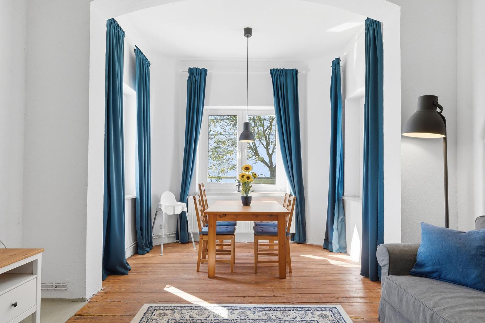 Dining area with wooden table, blue curtains, and sunflowers. Wooden floor and sofa in foreground.