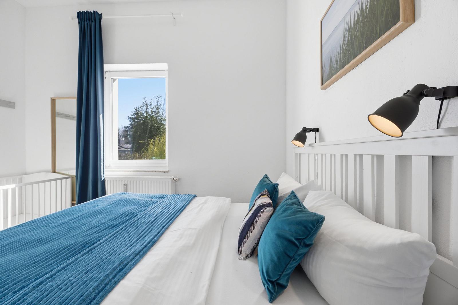 Bedroom with white bed, blue blanket, and wall lamp. Window with blue curtains.