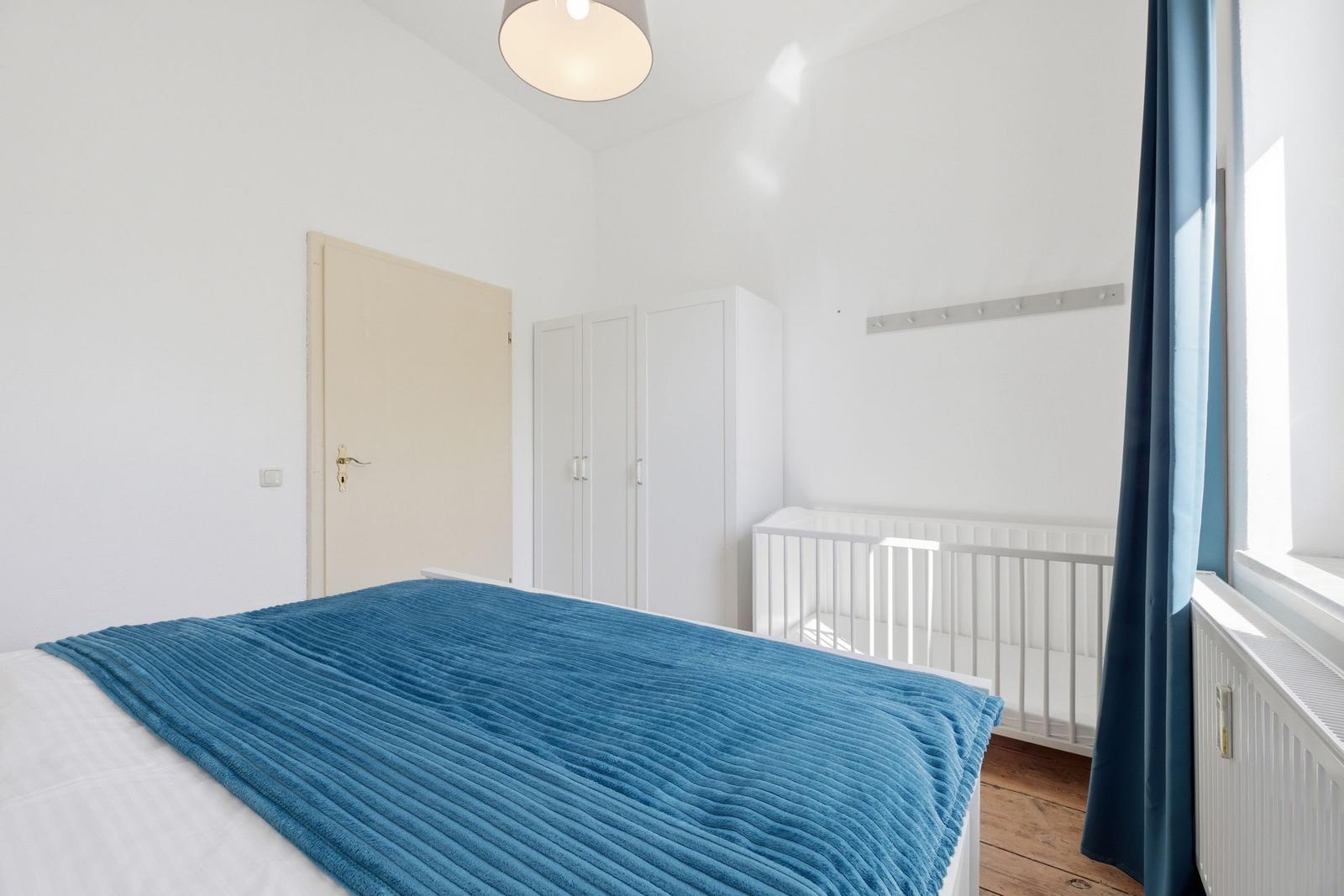 Bedroom with bed, crib, wardrobe, and window with curtain.