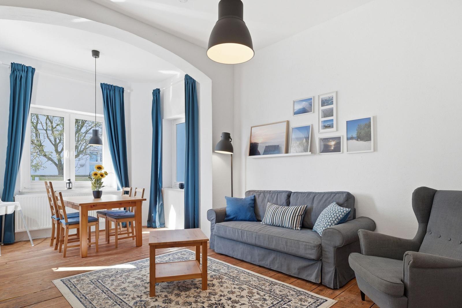 Living and dining area with sofa, chair, and table. Blue curtains and wall pictures.