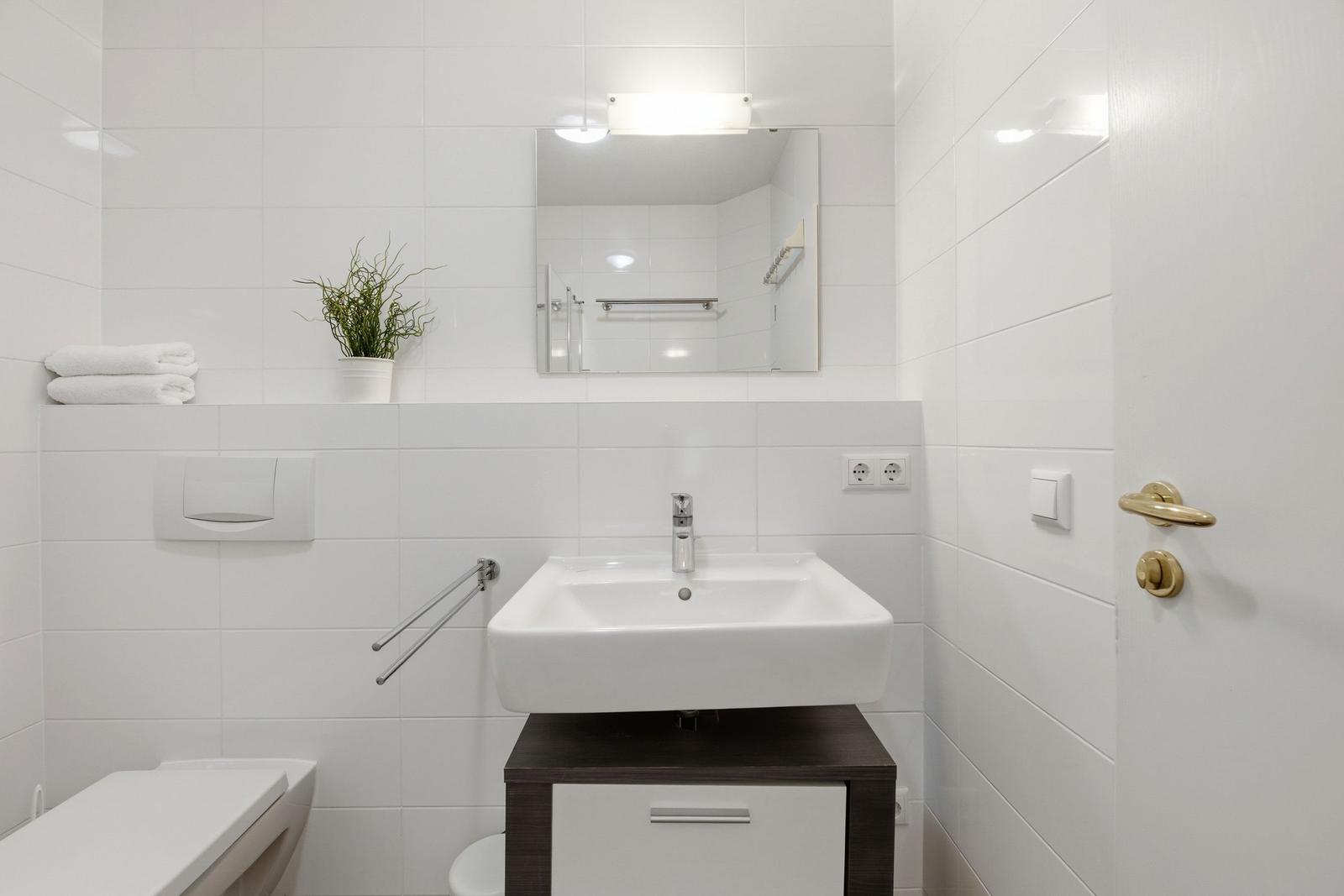 Badezimmer mit Waschbecken, Spiegel und Toilette. Weiße Fliesen und goldene Türknauf.
