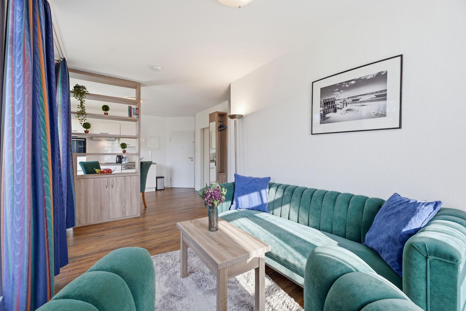 Ein Wohnzimmer mit grünem Sofa, Holztisch und Blick in die Küche.