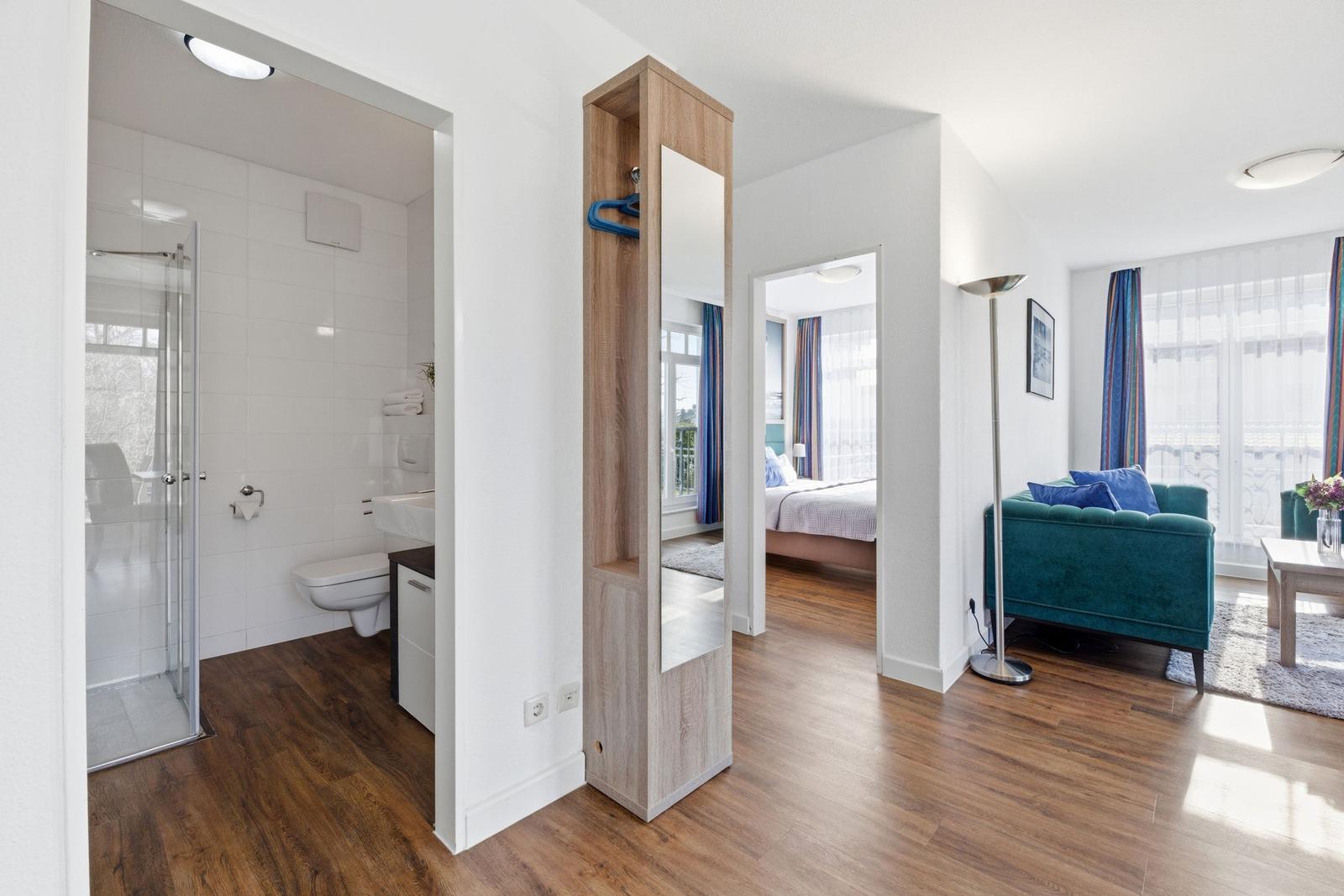 Badezimmer mit Dusche, Toilette und Waschbecken. Wohnzimmer mit Sofa und Schlafzimmer im Hintergrund.