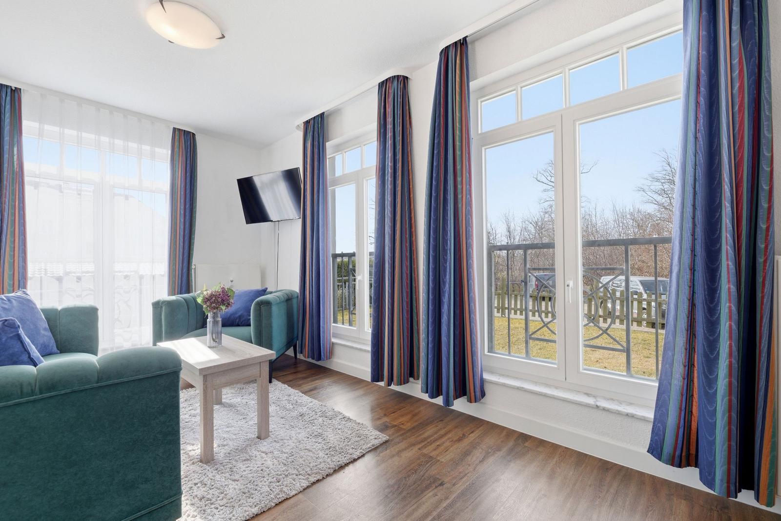 Wohnzimmer mit Sofa, Couchtisch und TV. Große Fenster mit Blick auf Balkon.