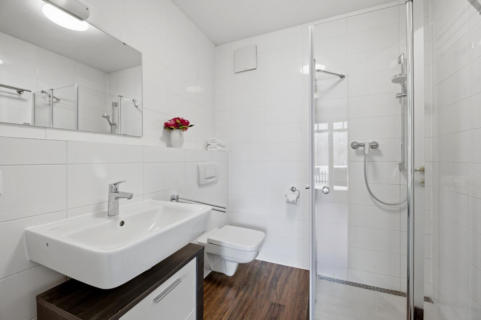 Badezimmer mit Dusche, Waschbecken und Toilette. Weiße Fliesen und Holzboden.