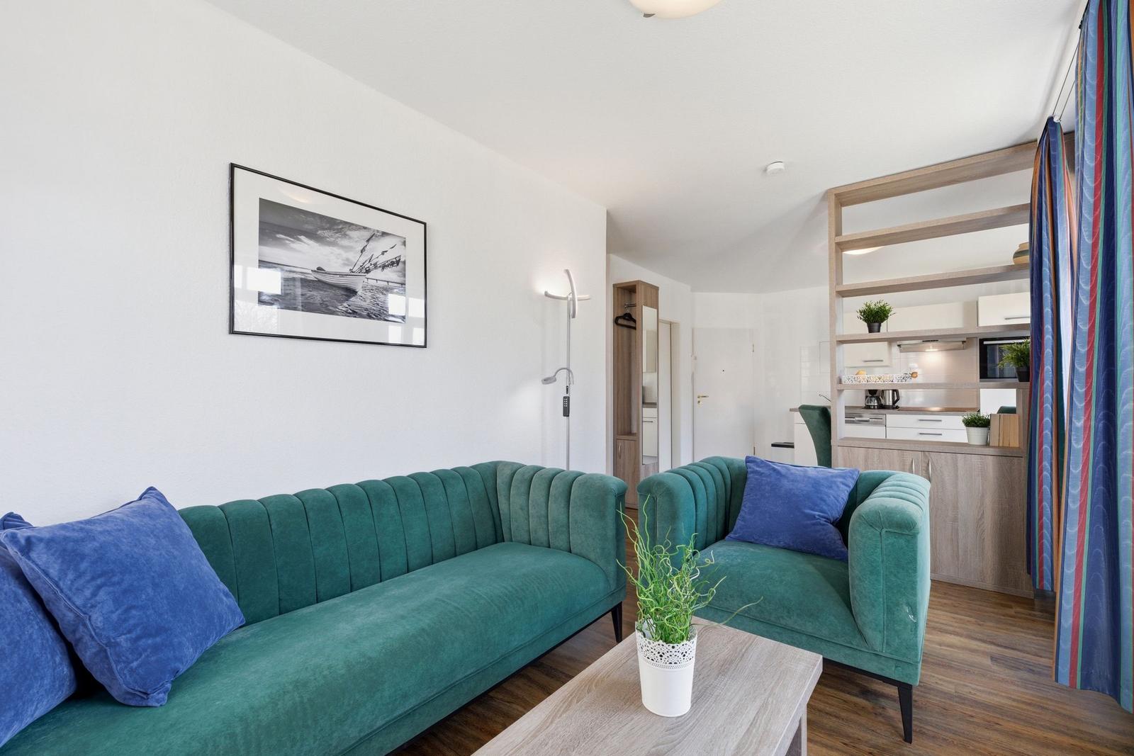 Ein Wohnzimmer mit grünem Sofa, blauen Kissen und einem Holztisch. Im Hintergrund ist die Küche sichtbar.