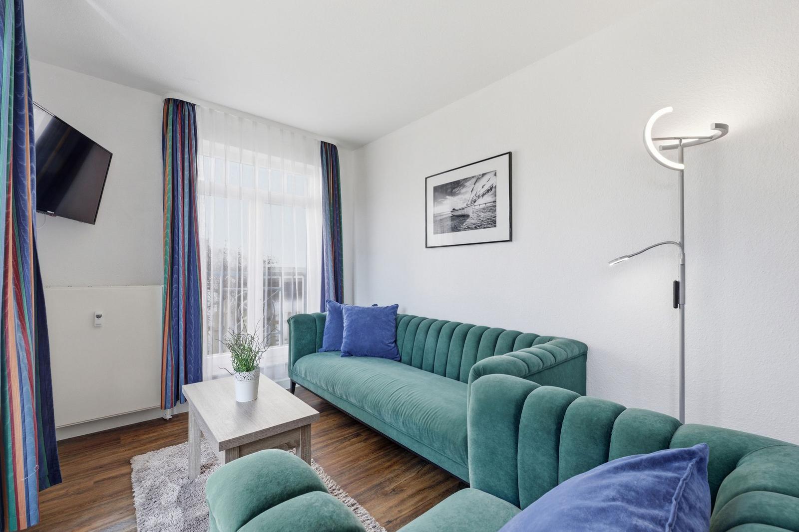 Ein Wohnzimmer mit grünem Sofa, blauen Kissen und einem Fernseher an der Wand.