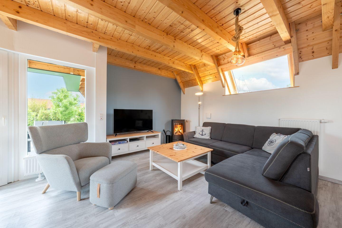 Wohnzimmer mit Holzdecke, Sofa, Sessel und Kaminofen. Fenster und Dachfenster bieten Blick auf Außenbereich.