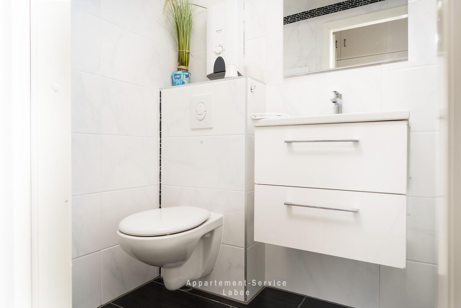 Badezimmer mit Toilette, Waschbecken und Spiegel. Weiße Fliesen und dunkle Bodenfliesen.