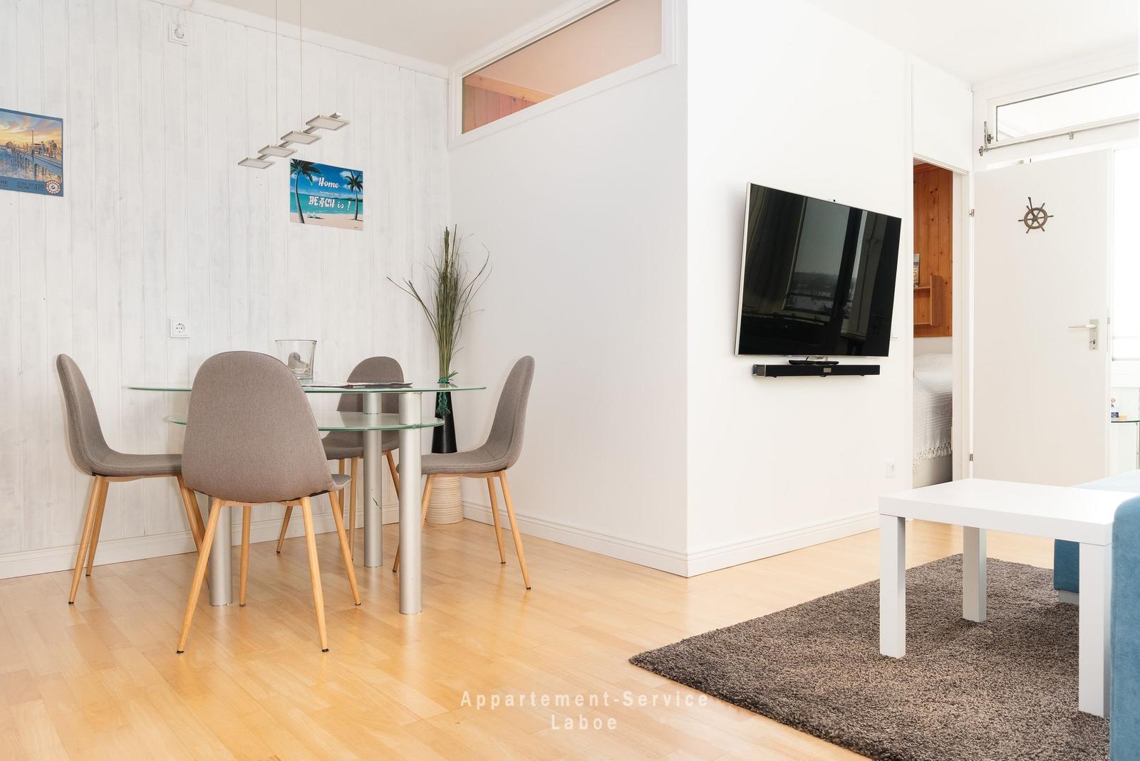Essbereich mit Glasplatte und grauen Stühlen. Fernseher an Wand. Tür zum Schlafzimmer.