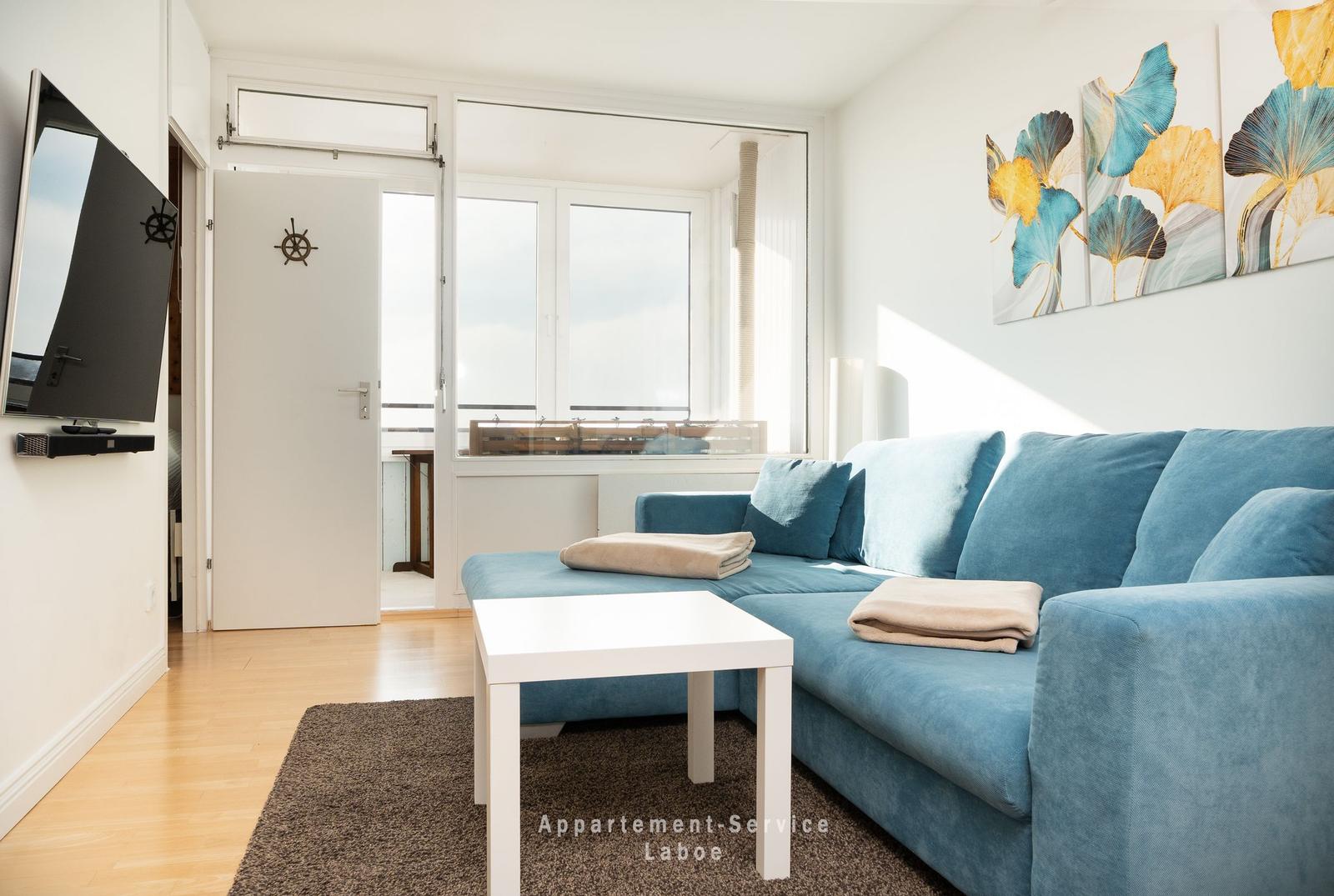 Blauer Sofa, weißer Couchtisch, Fernseher an Wand, Fenster mit Blick auf Balkon.