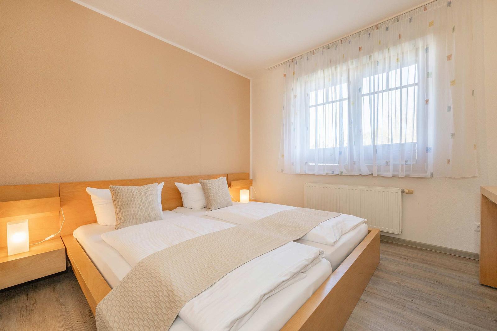 Schlafzimmer mit Doppelbett, Holzgestell, Fenster mit Vorhängen und Nachttisch mit Lampe.