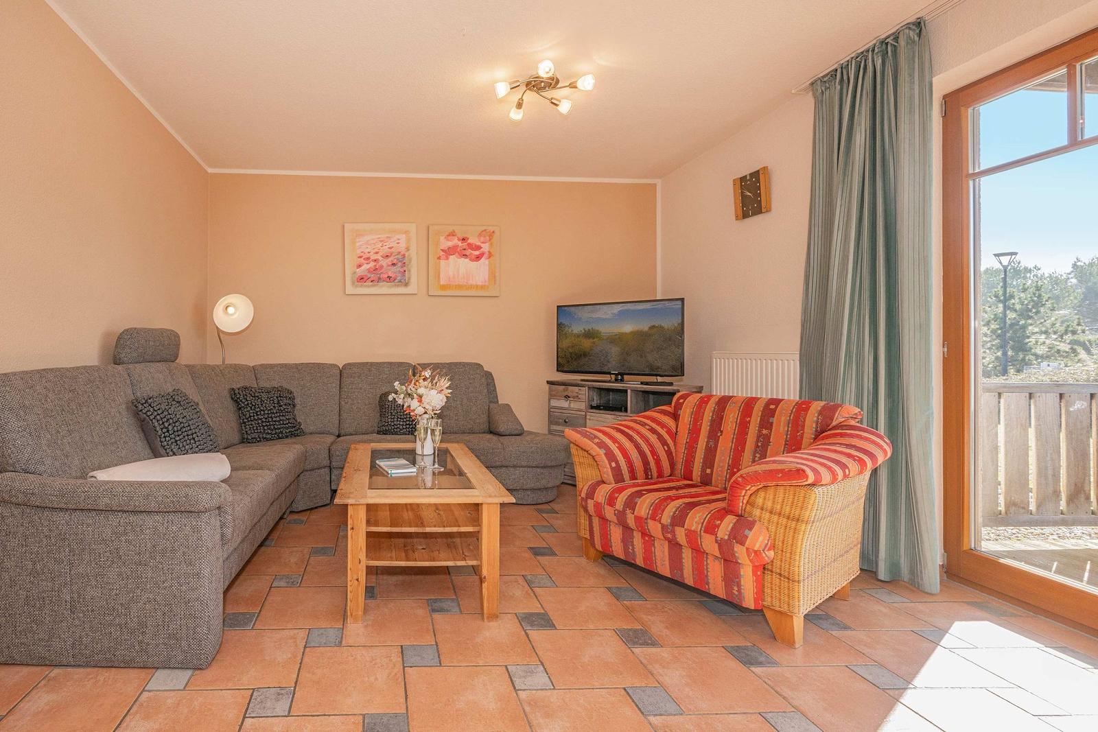 Wohnzimmer mit Sofa, Sessel, Couchtisch und Fernseher. Terrasse mit Blick auf Garten.
