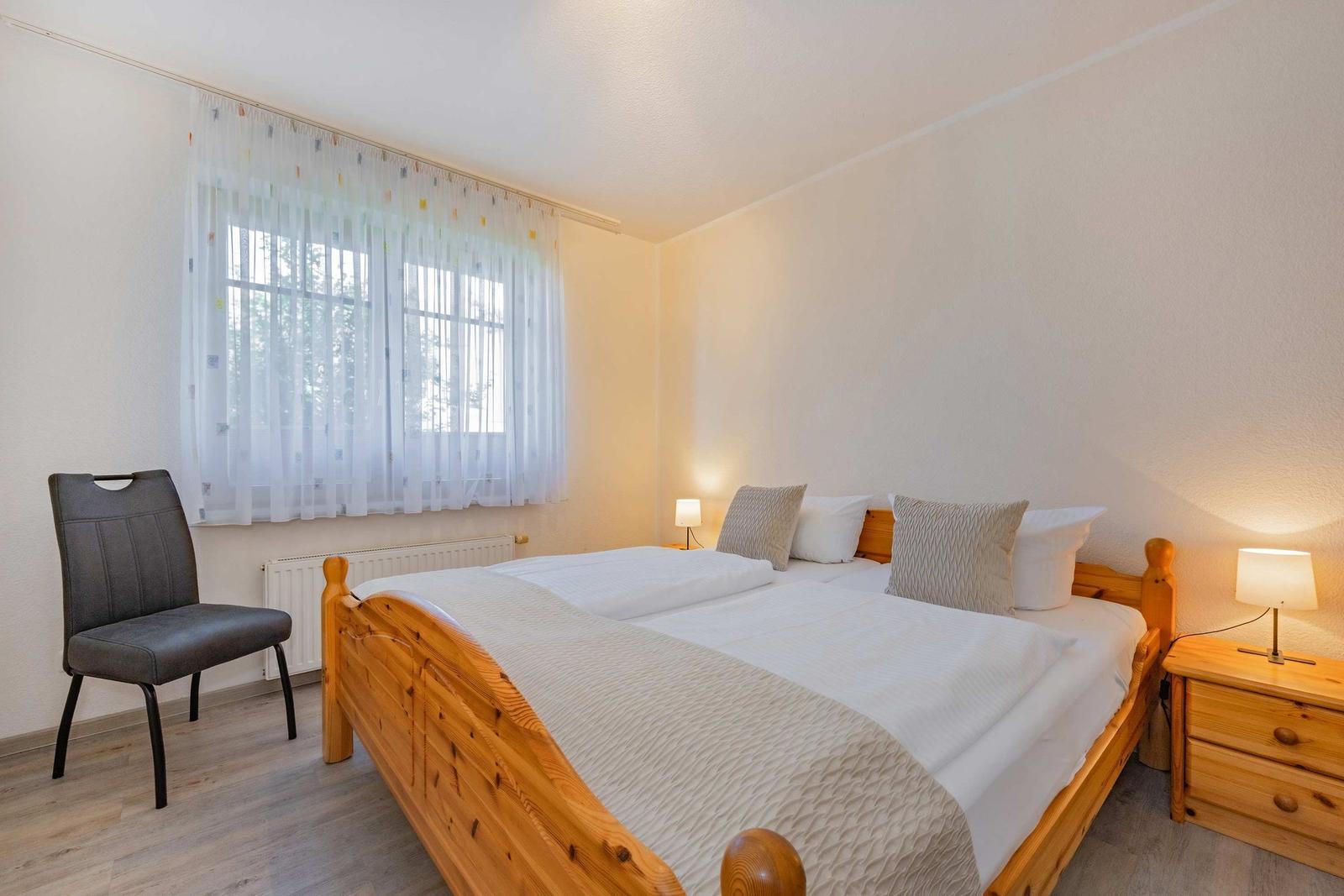 Schlafzimmer mit Holzbett, Nachttisch, Stuhl und Fenster mit Vorhängen.