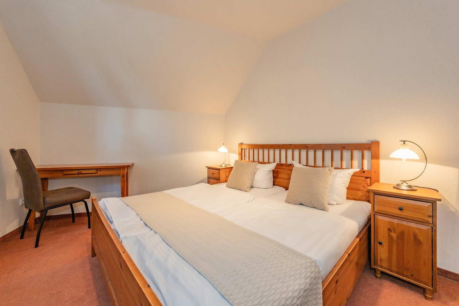 Schlafzimmer mit Holzbett, Nachttischen und Schreibtisch unter schrägem Dach.