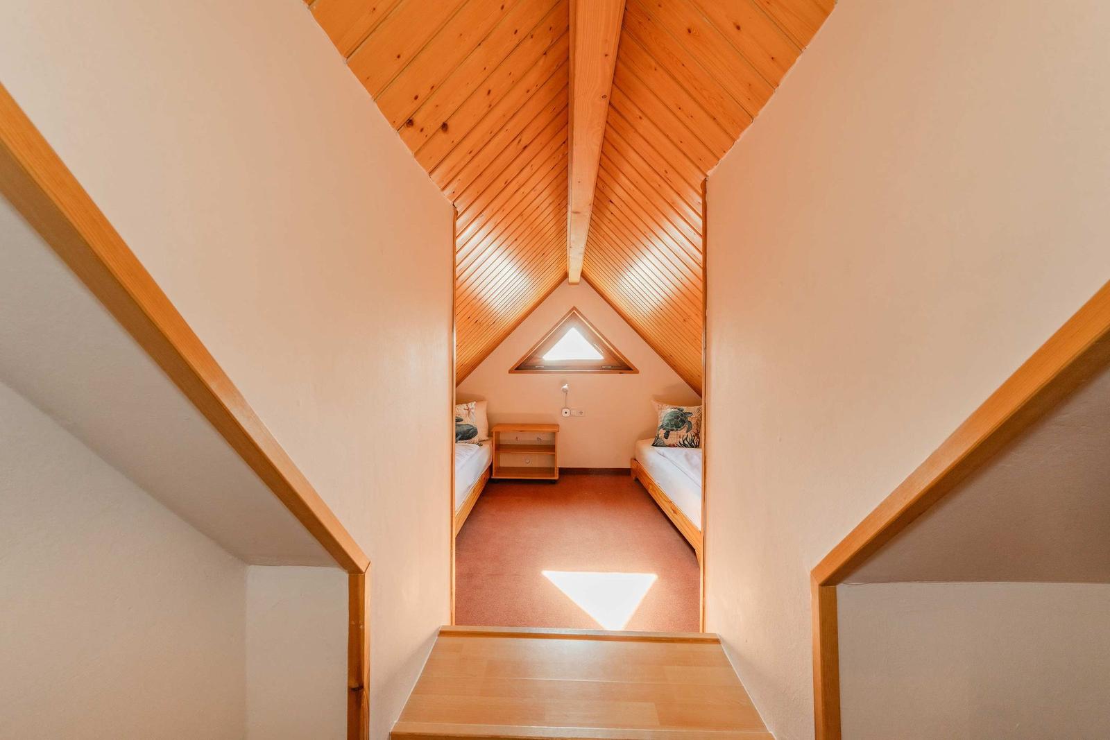 Zimmer mit zwei Betten unter Holzdach, kleiner Schrank und Dreiecksfenster.