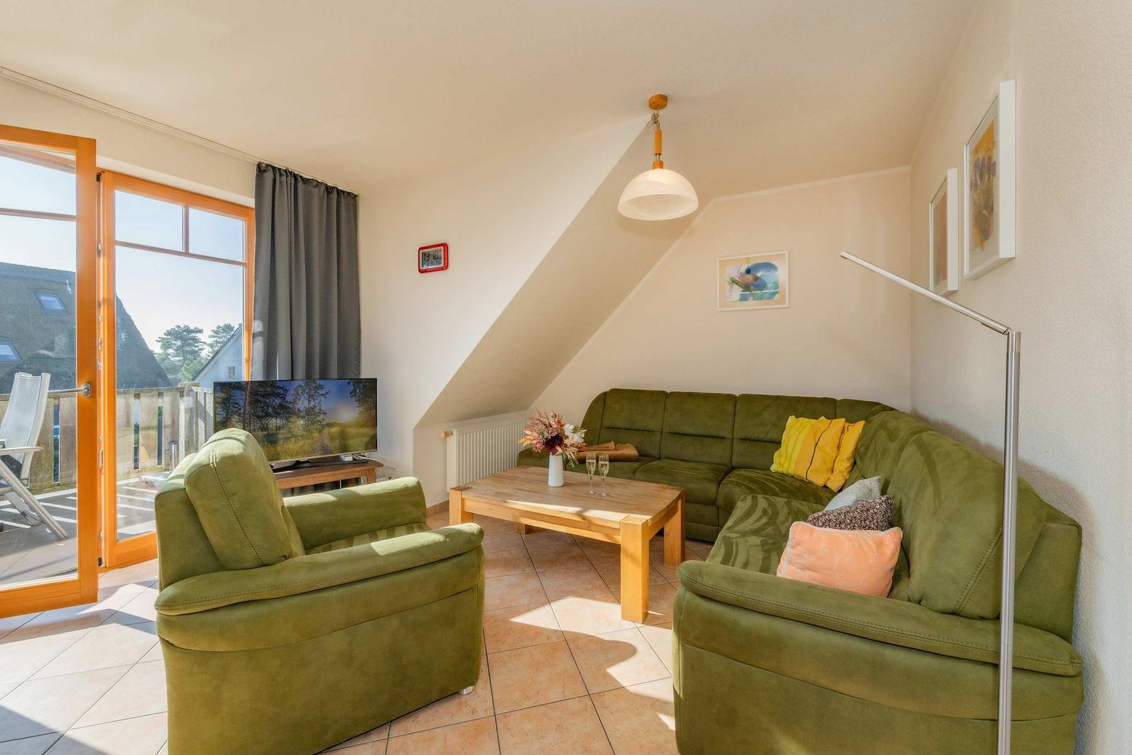 Wohnzimmer mit grünem Sofa, Holztisch und TV. Durchgang zur Terrasse mit Blick auf Garten.