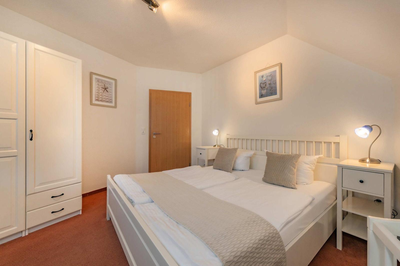 Schlafzimmer mit Doppelbett, Nachttisch und Kleiderschrank. Wandbild und Deckenleuchte sichtbar.