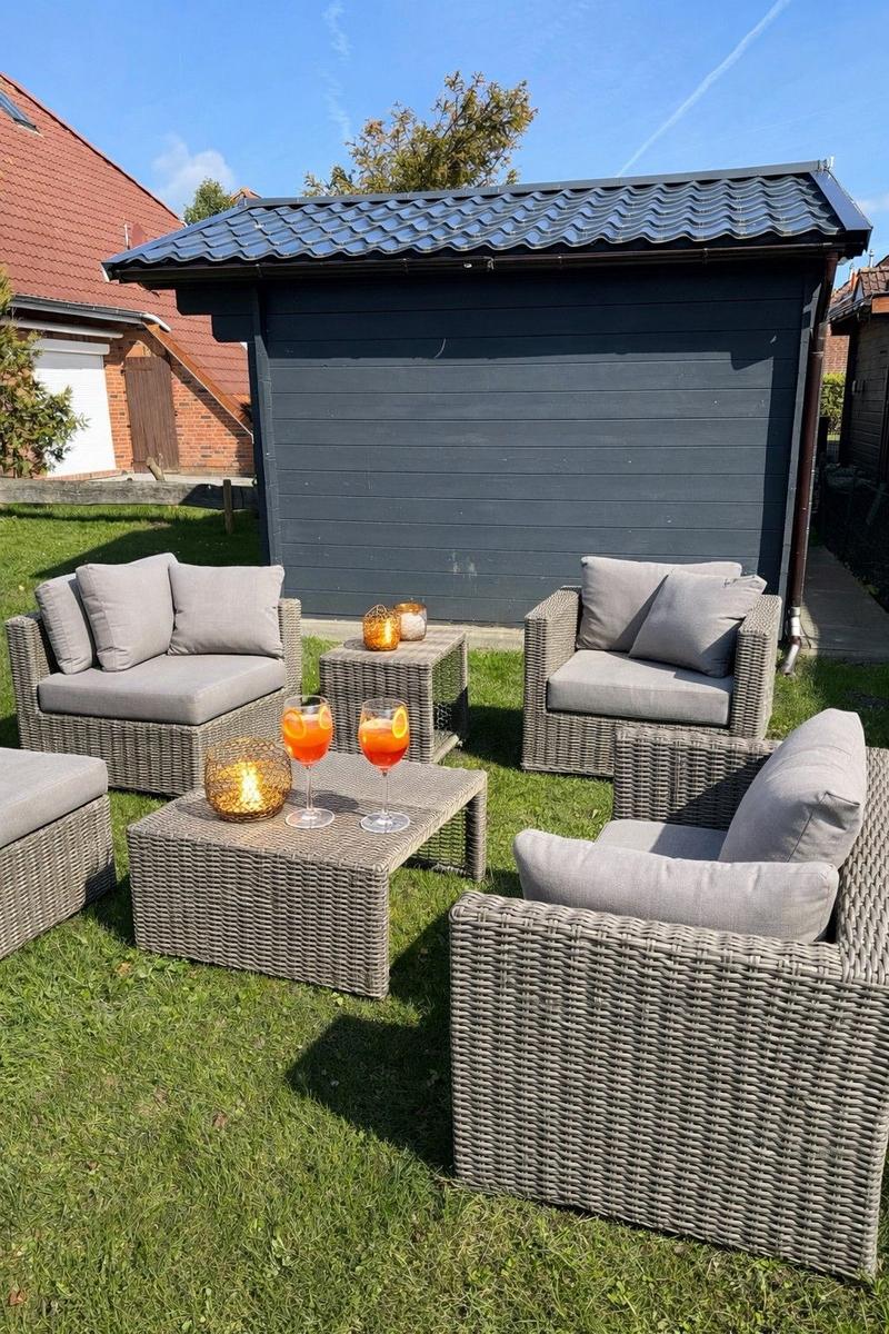 Gartenbereich mit Rattanmöbeln und Getränken vor einem grauen Gartenhaus.