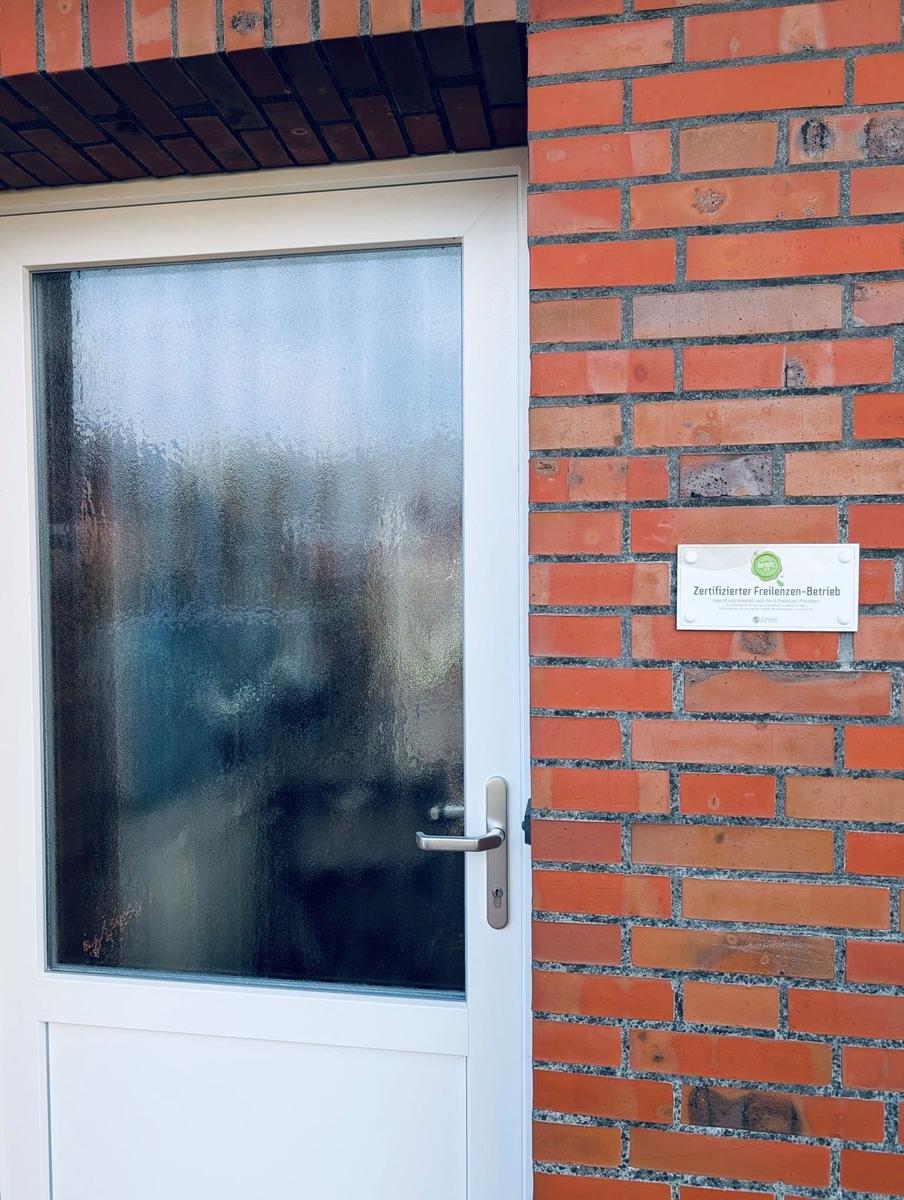 Eingangstür mit Glas und Schild Zertifizierter Freienzen-Betrieb an rotem Ziegelsteinhaus.