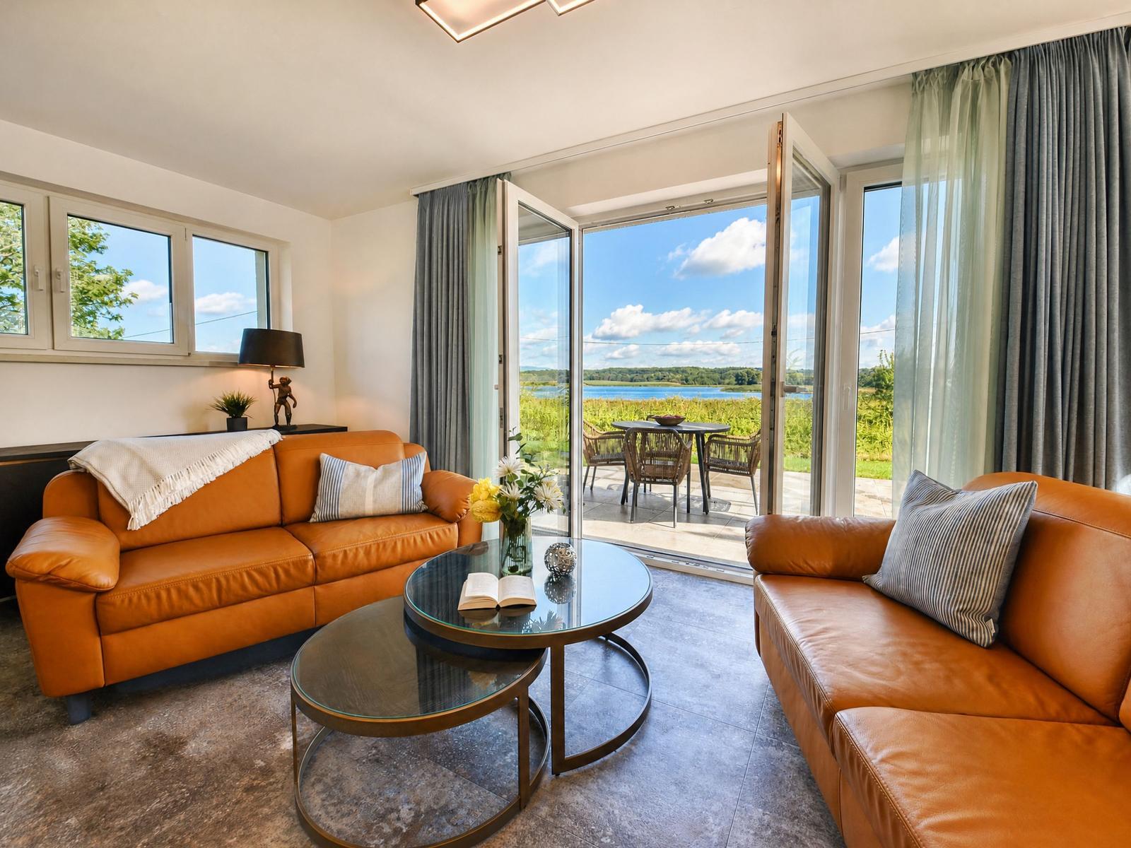 Wohnzimmer mit braunen Sofas, Glas-Tisch und Blick auf den See.