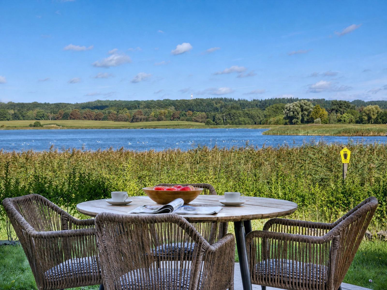 Rattan-Tisch mit Stühlen am See, Obstschale, Tasse und Zeitung. Blick auf Wald und Wasser.
