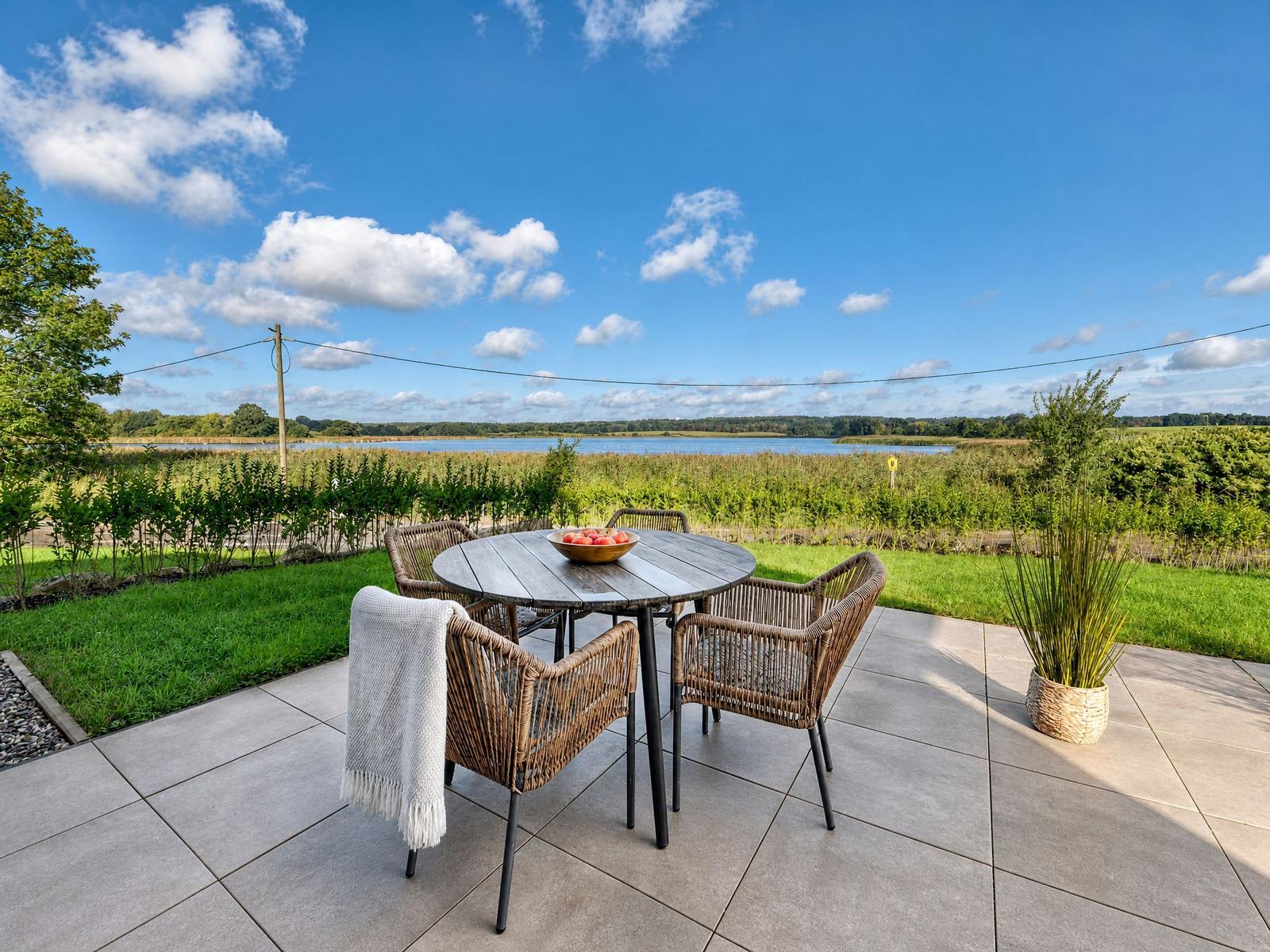 Terrasse mit runden Tisch und Stühlen, Blick auf Wasser und Grünflächen.