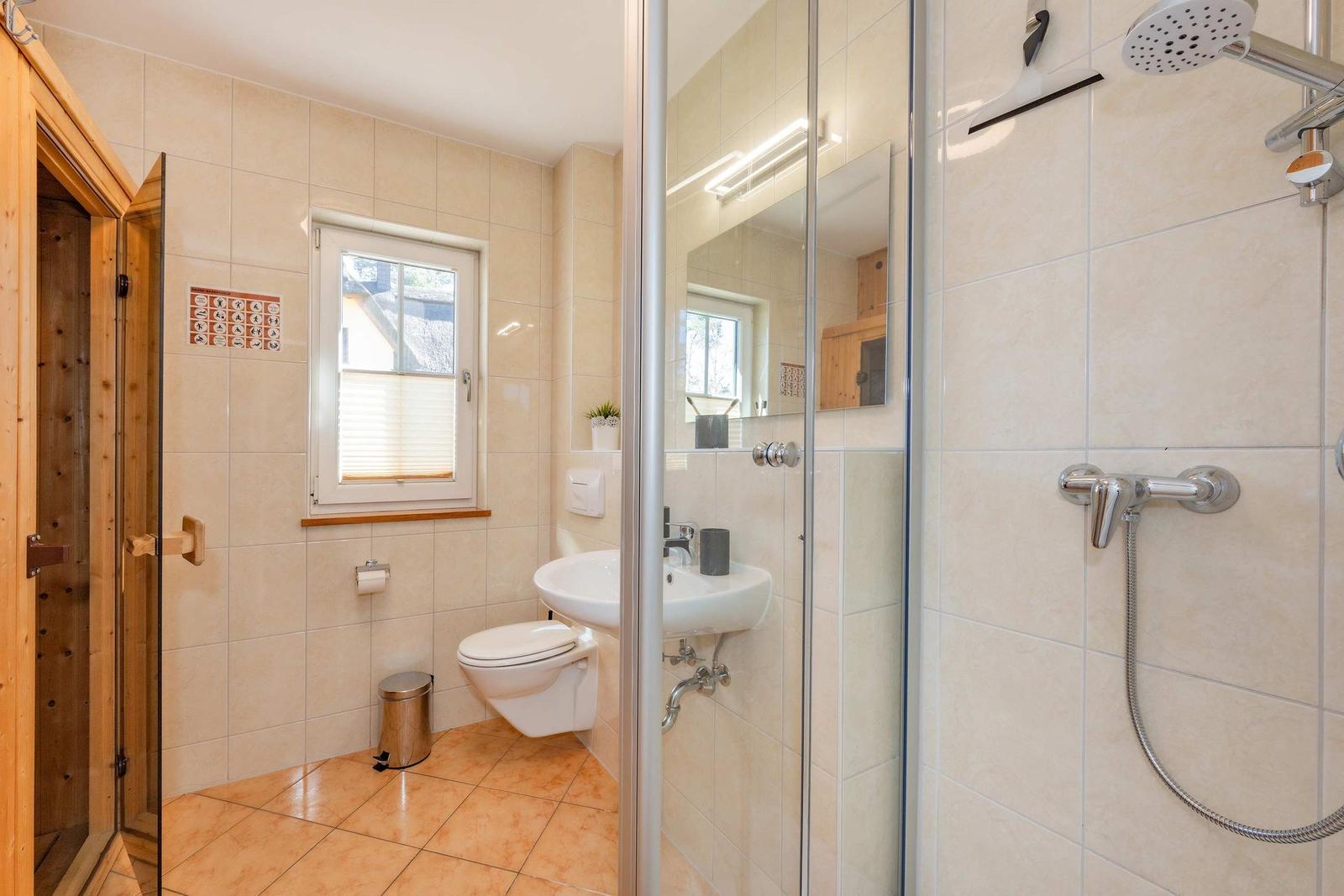 Badezimmer mit Dusche, Toilette und Waschbecken. Holzverkleidung und Fenster mit Jalousie.