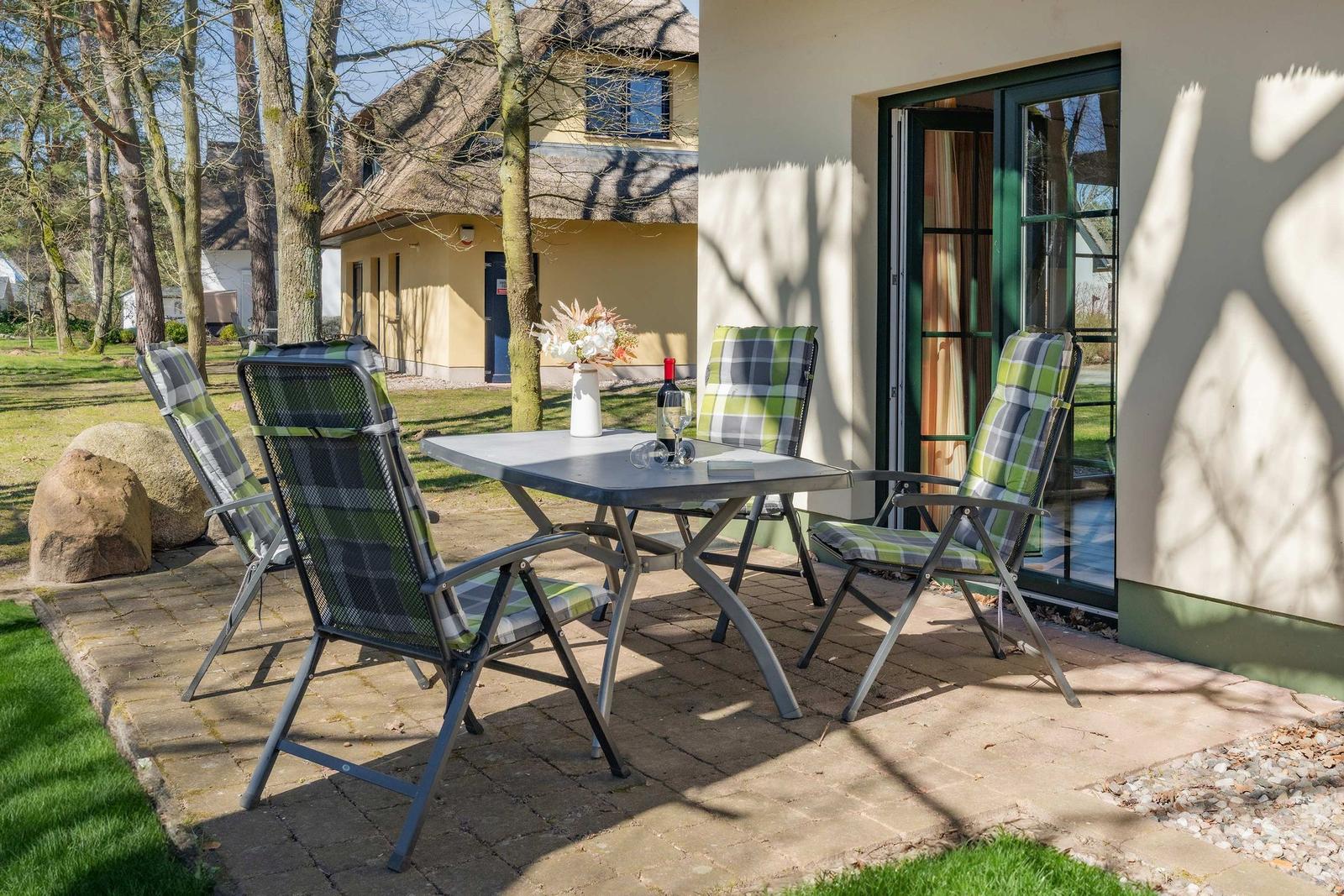 Terrasse mit Tisch und vier Stühlen, daneben Haus mit Glastür und Garten.