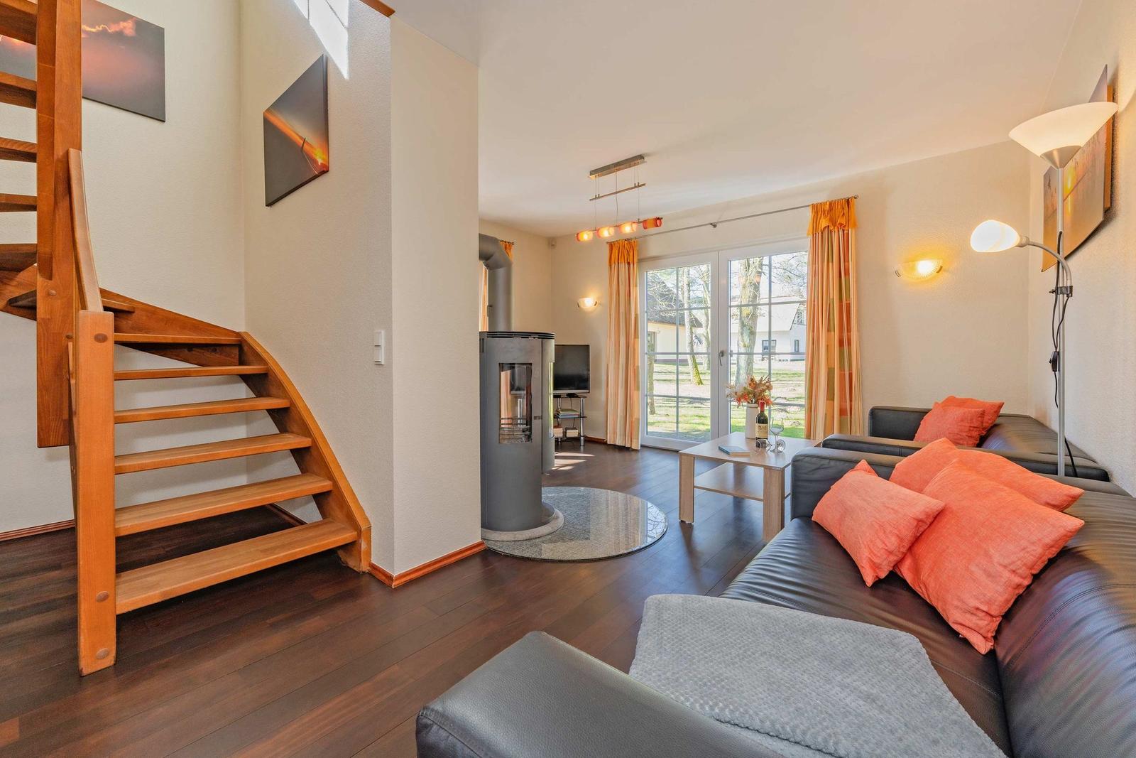 Wohnzimmer mit Sofa, Kamin und Holztreppe. Fenster mit Vorhängen zur Außenterrasse.