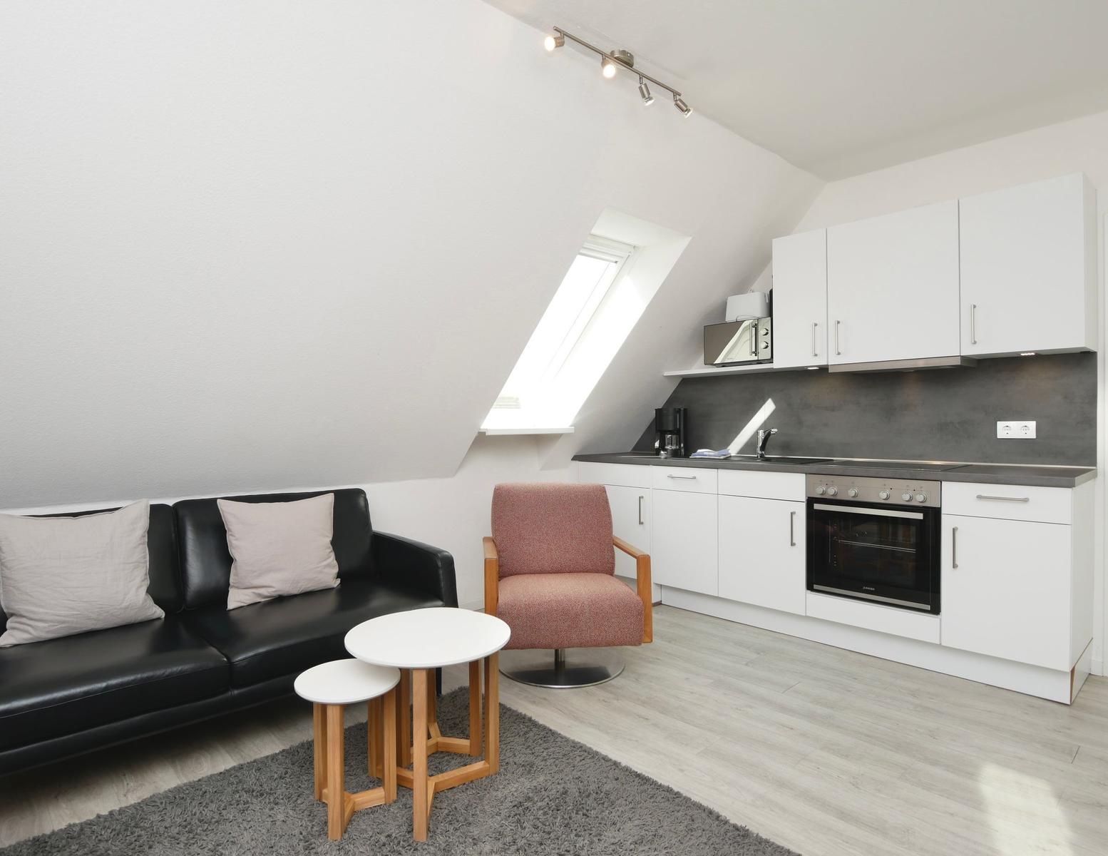 Wohnbereich mit Sofa, Sessel und Küchenzeile unter Dachschrägen.