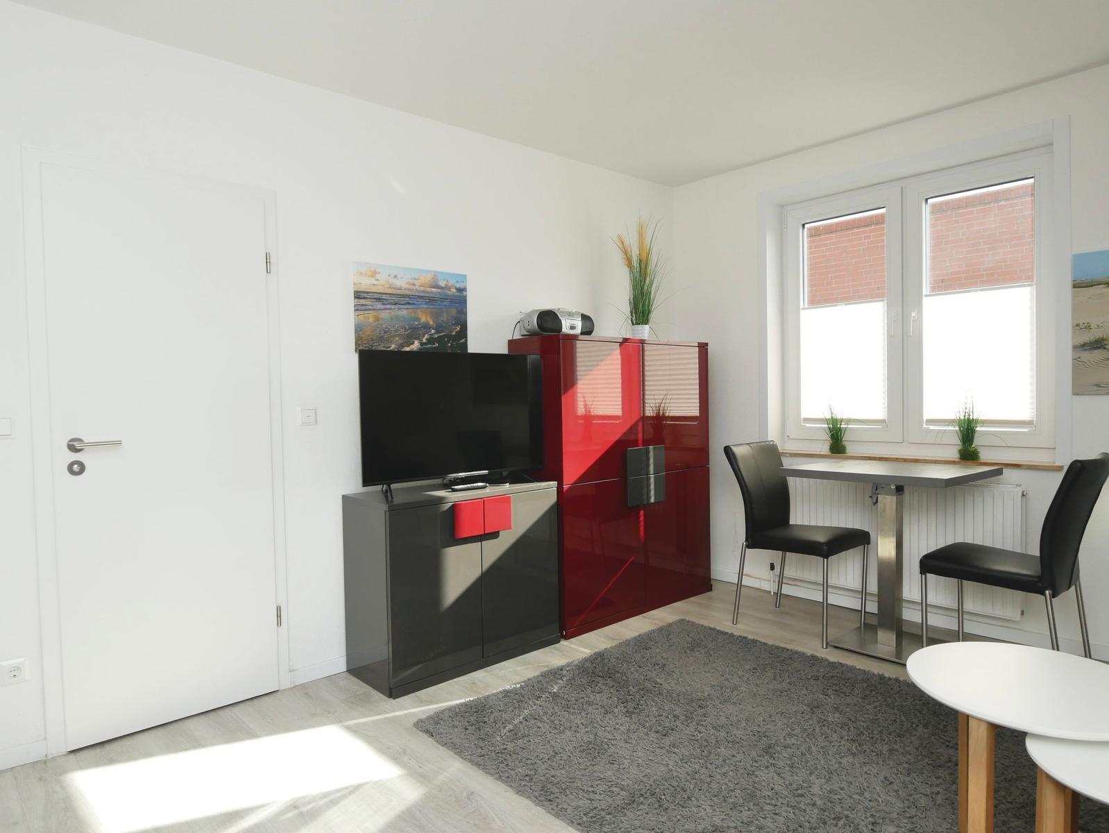 Wohnzimmer mit TV, rotem Schrank und Esstisch. Fenster mit Blick auf Backsteinmauer.