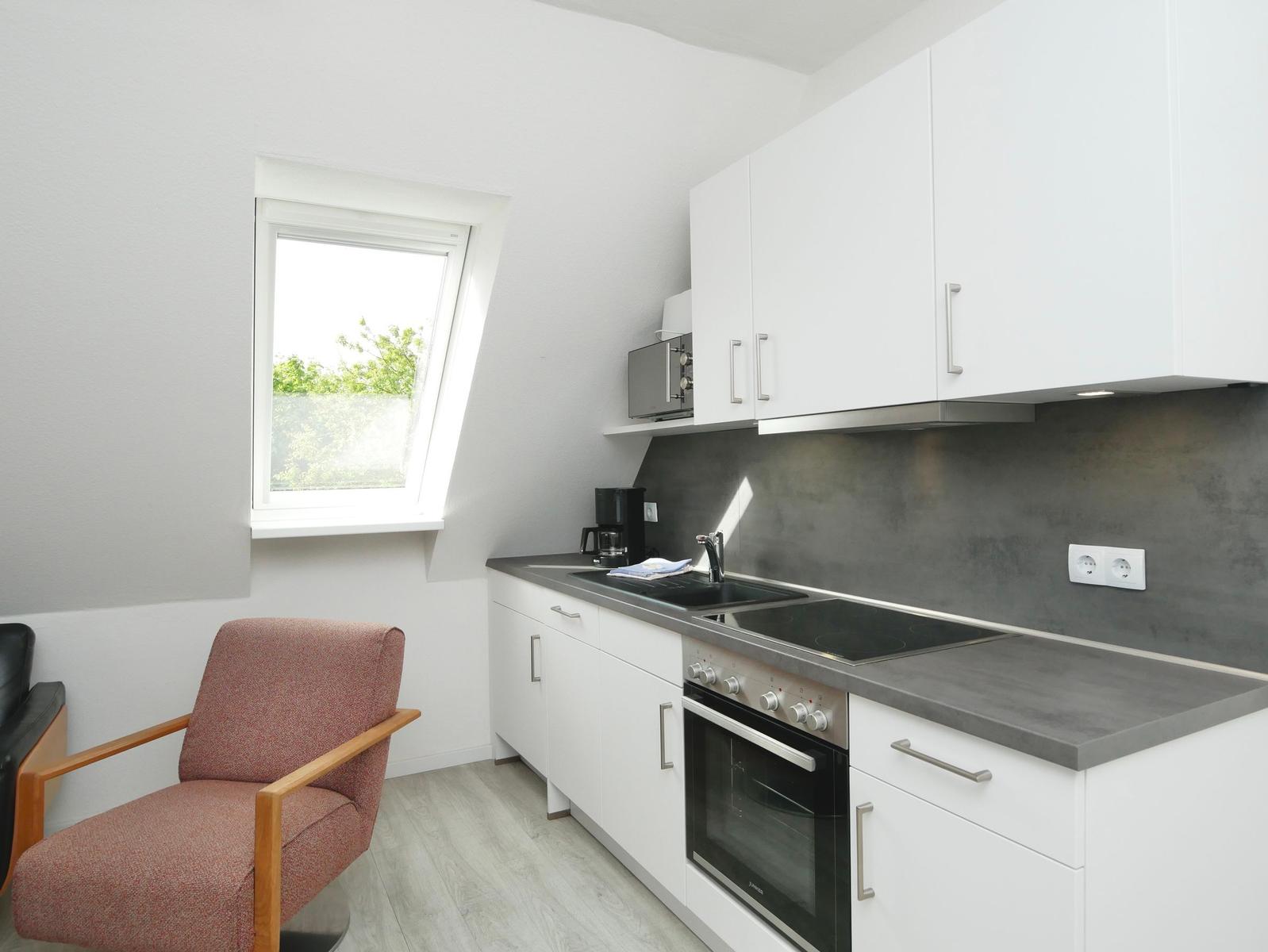Küche mit weißer Einbauküche, grauer Arbeitsplatte und Fenster. Ein roter Sessel steht daneben.