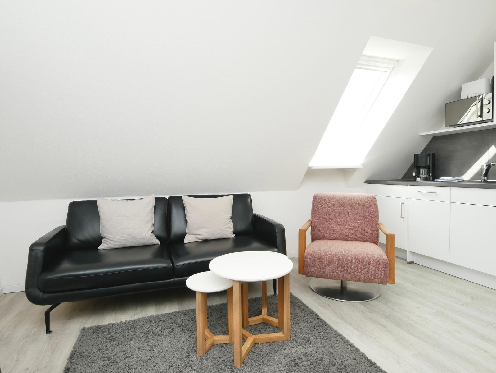 Schwarzes Sofa mit Kissen, rosa Sessel und Küchenzeile unter Dachschrägen.