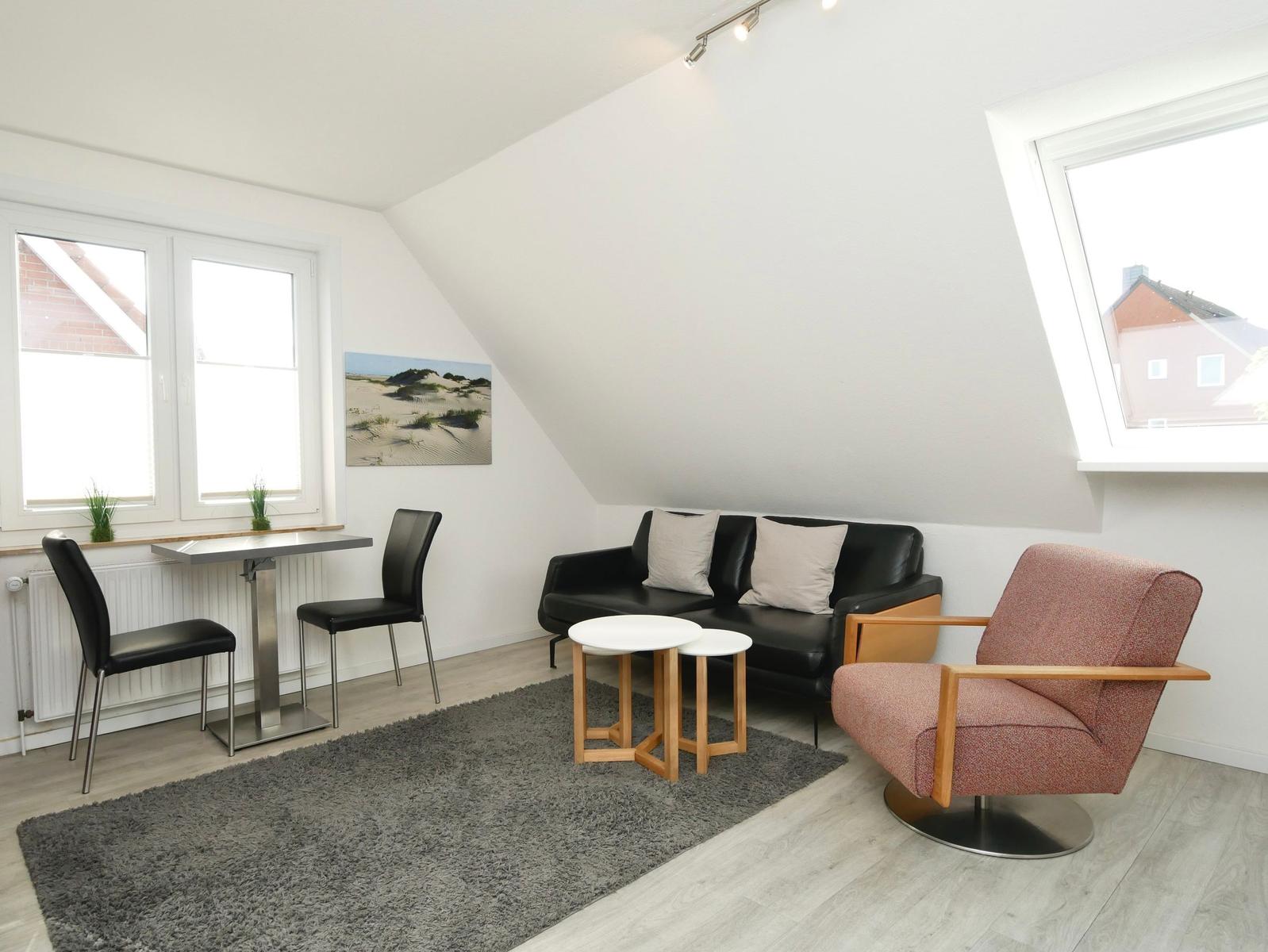 Wohnraum mit Sofa, Sessel, Tisch und Fenstern. Dachschrägen und Teppichboden.