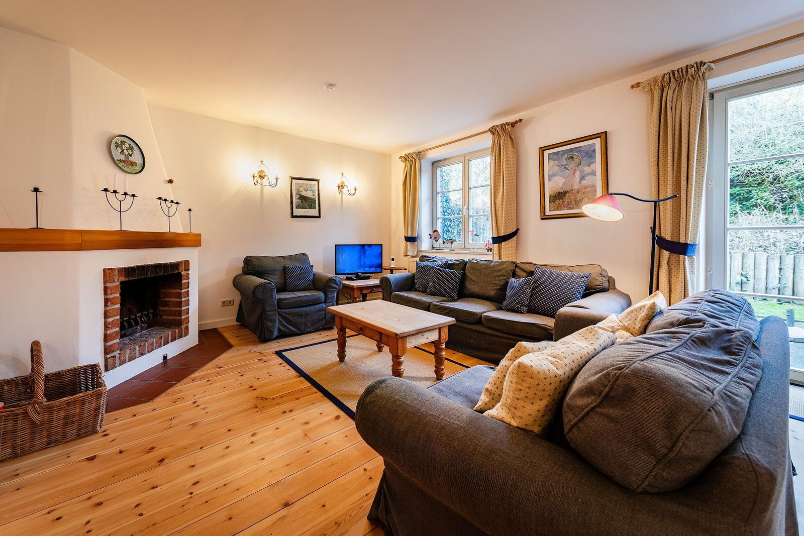 Wohnzimmer mit Kamin, Sofa, Sessel und Holzboden. Fenster mit Vorhängen und Fernseher.