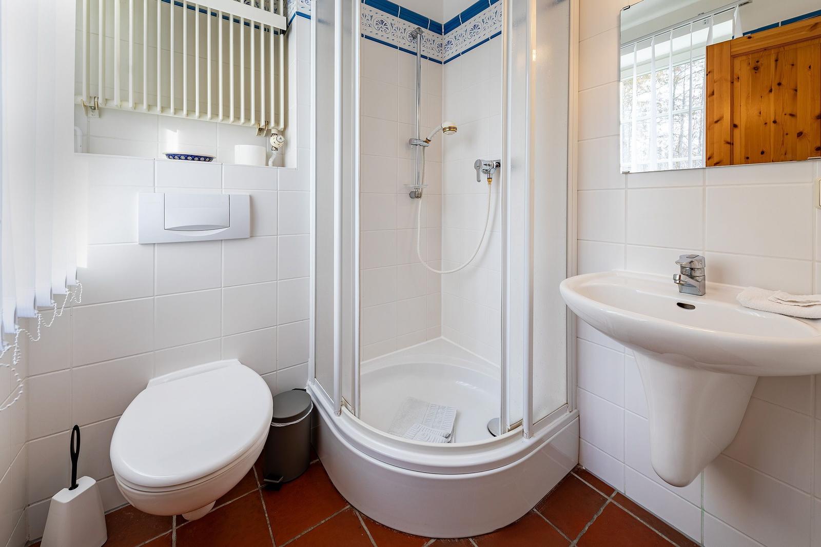 Badezimmer mit Dusche, Waschbecken und Toilette. Weiße Fliesen und Terrakottaboden.